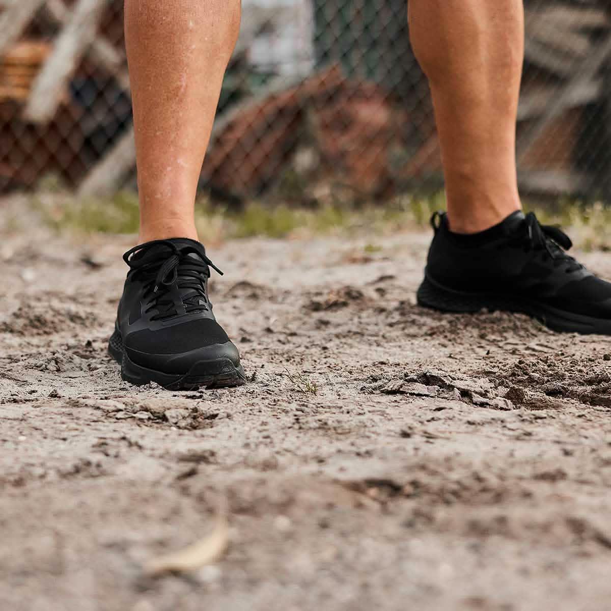 A person wearing GORUCKs Rough Runner - Blackout stands on a dirt path with rocks and grass in the background. The image focuses on the shoes and lower legs, suggesting a Rough Runner event or sporty setting.