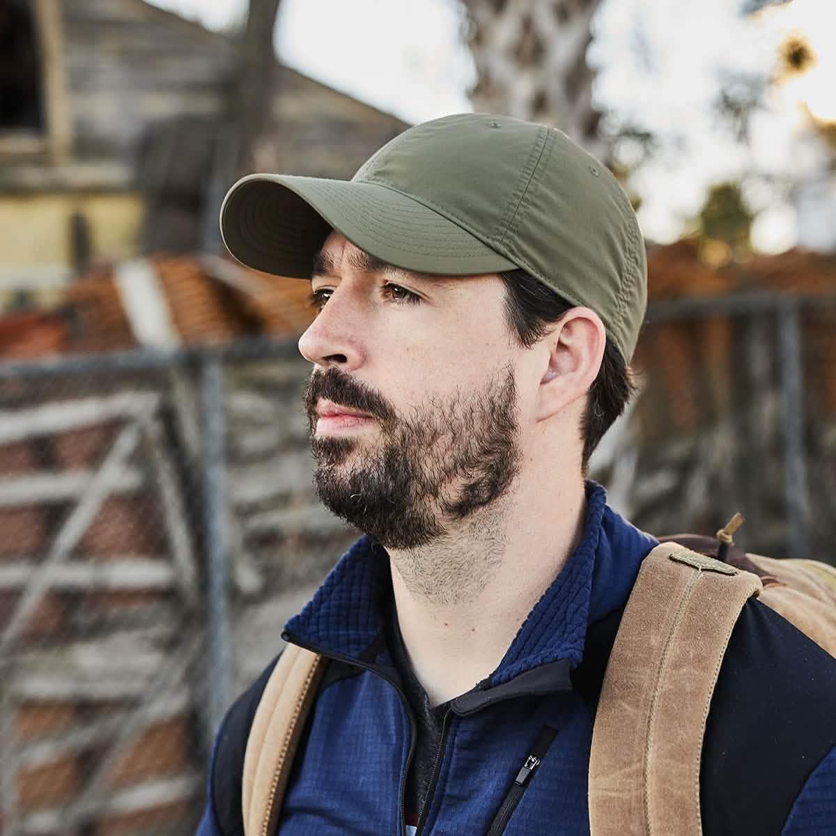 Man wearing olive GORUCK cap and tan rucksack outdoors, rugged background