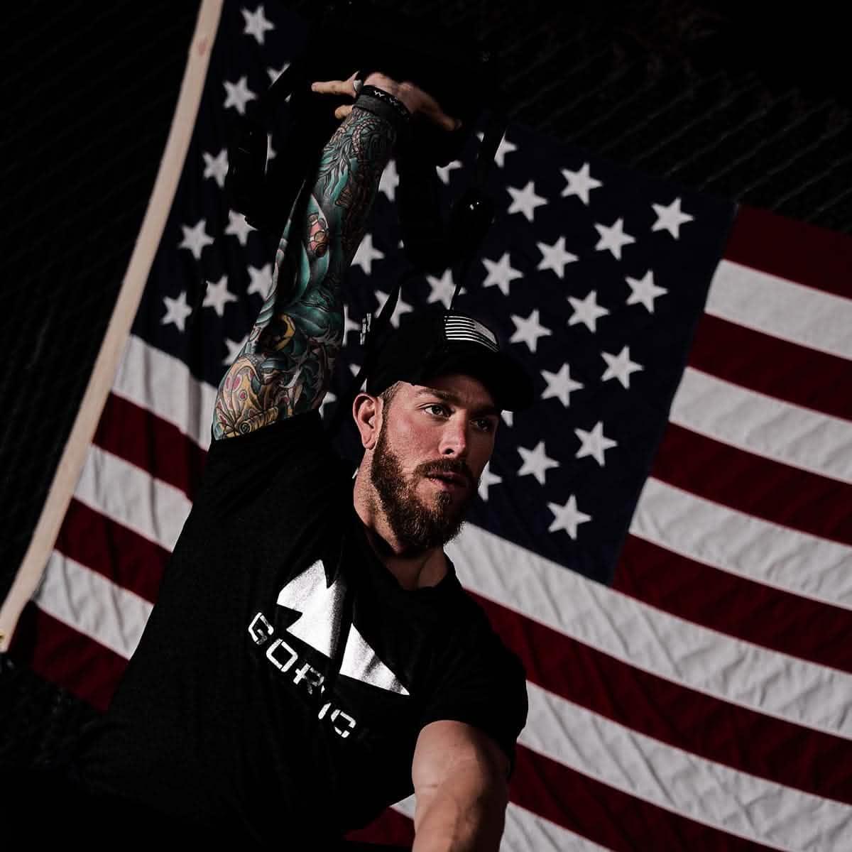 Man lifting weight wearing GORUCK reflective tee and hat, American flag in background