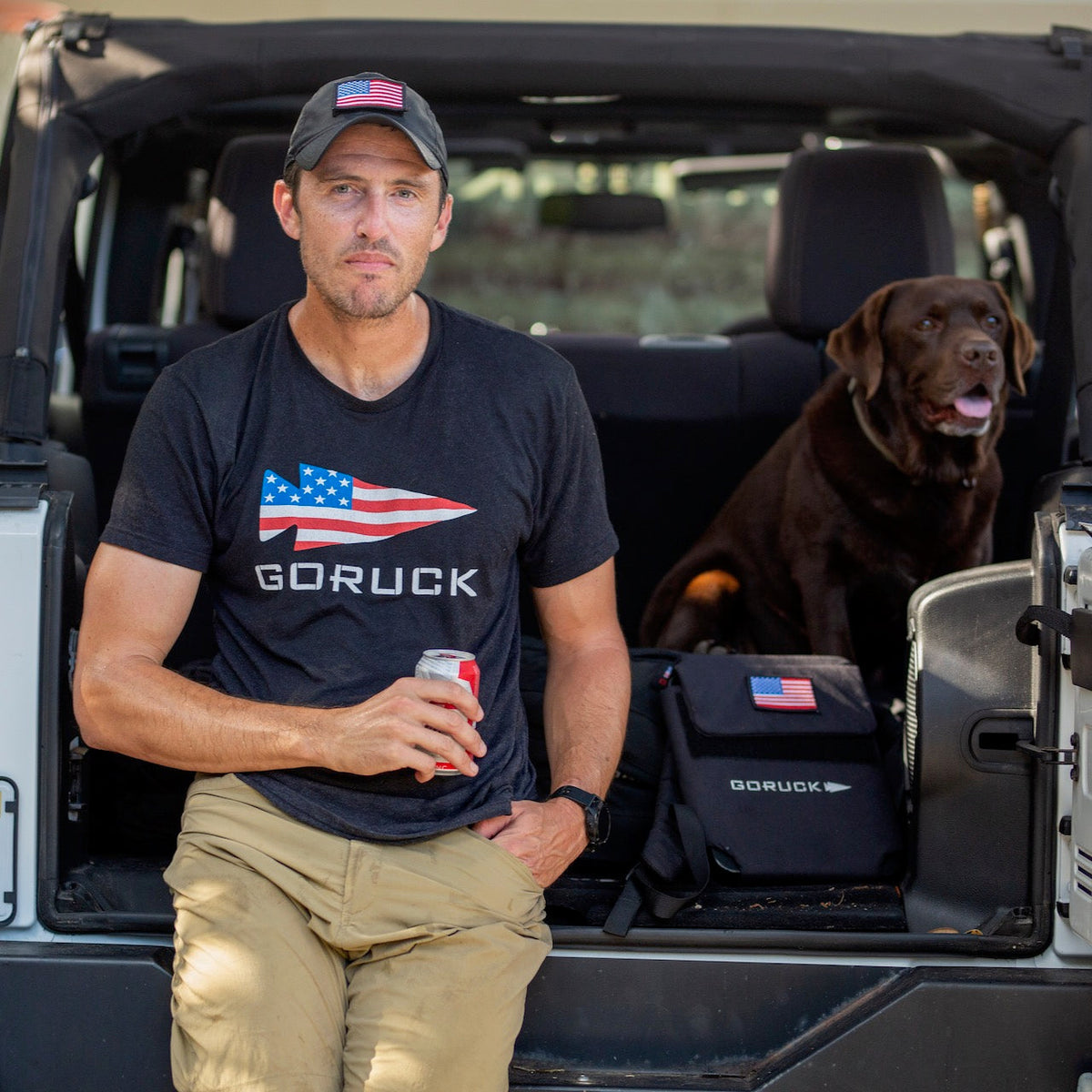 A man in a GORUCK shirt sits on a car tailgate with his brown dog and a Ruck Plate Carrier 3.0 weighted backpack, enjoying a soda outdoors.