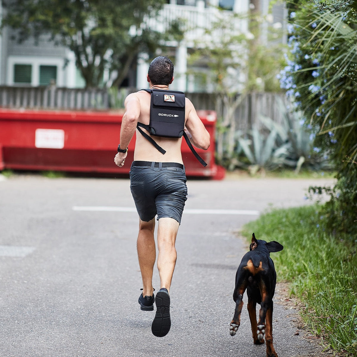 A shirtless man runs with a backpack down the street, followed by a black dog, showcasing the Spearhead Nylon Webbing Belt—an adjustable belt designed for both comfort and durability.