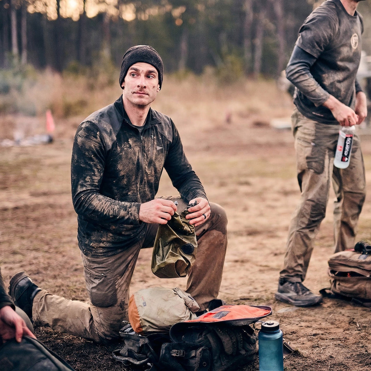 A man kneels on muddy ground holding a cap, while another stands nearby; both wear dirty outdoor gear, including the Men’s Commando Half Zip - Merino Wool sweater with reinforced shoulders for extra durability.