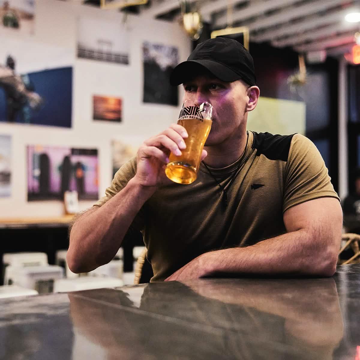 Man in GORUCK apparel wearing a black cap, drinking beer at a bar with photo wall decor.
