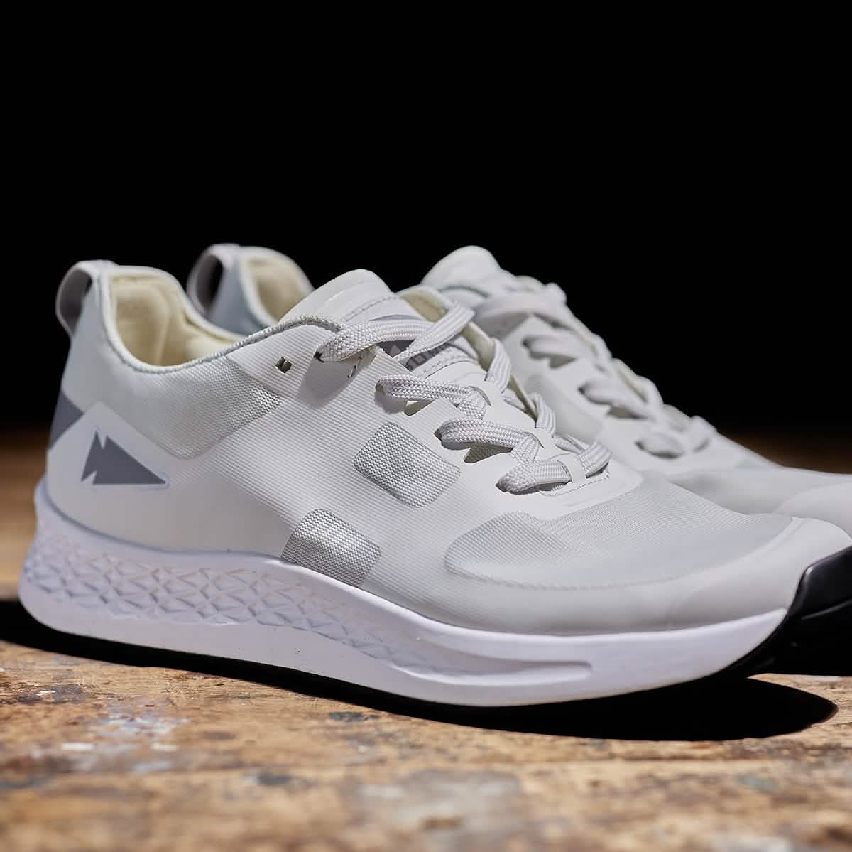 A pair of GORUCK Rough Runner sneakers in light grey and dark grey, featuring a white gradient density sole and a triangular logo on the side, are placed on a wooden surface. The shoes include laces and a heel loop for easy wearing, with the dark background providing the perfect contrast to showcase these sneakers.