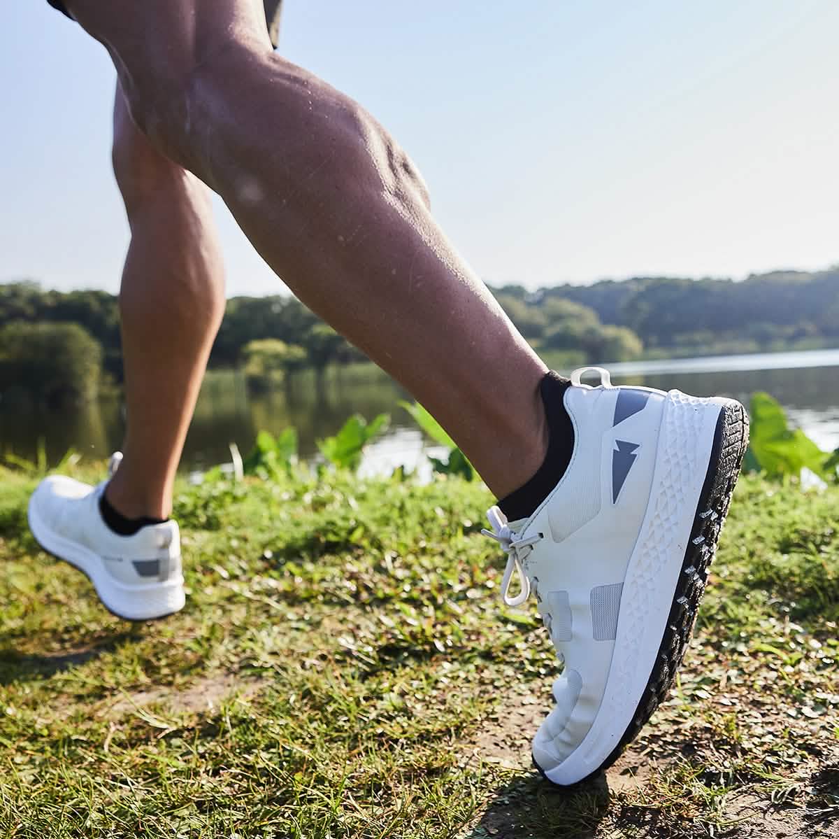 A person jogging outdoors next to a lake embodies the spirit of GORUCK's Rough Runner shoes in Light Grey + Dark Grey. The focus is on their legs and athletic footwear, set against a backdrop of green grass and trees under a clear blue sky.
