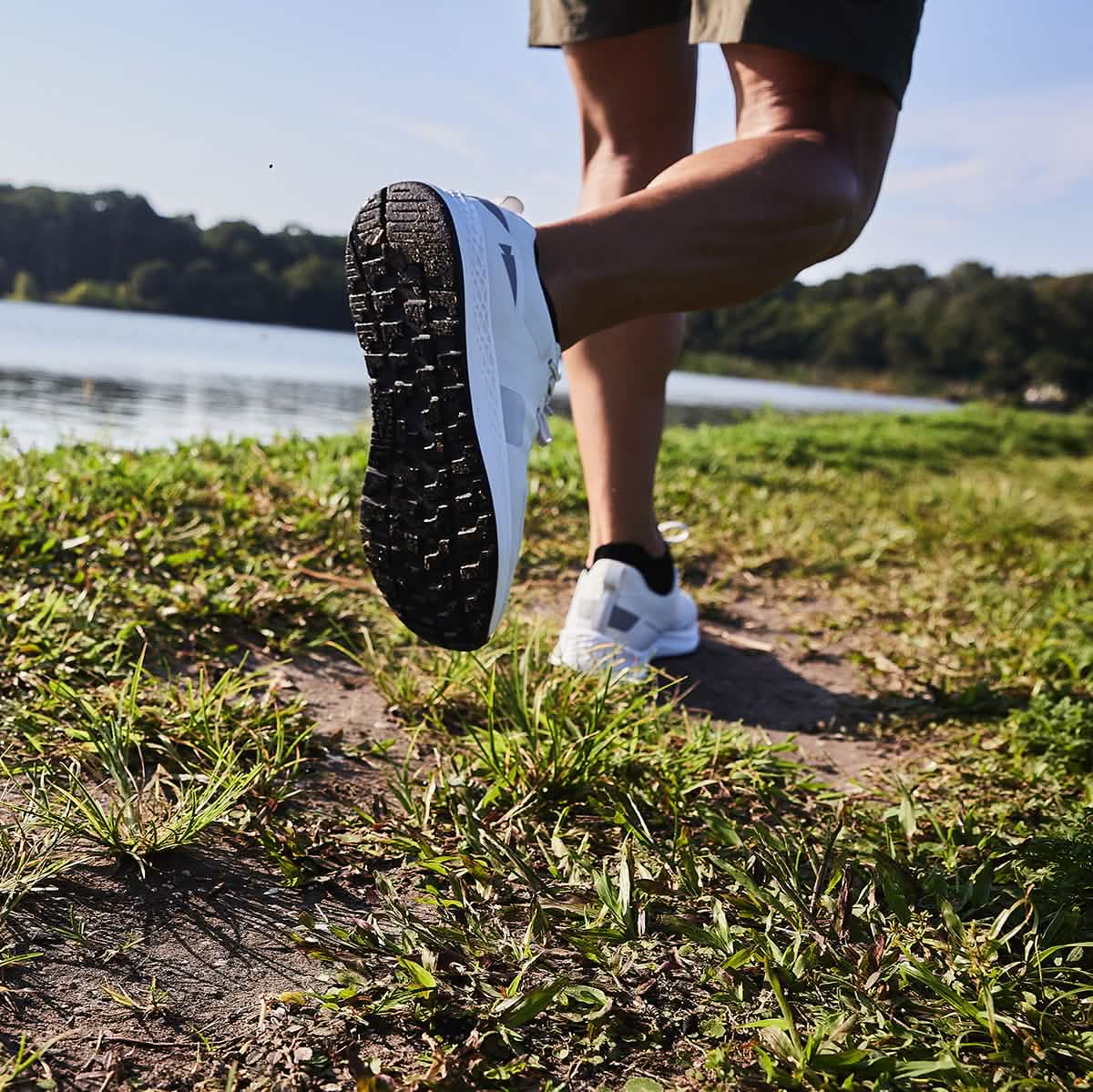 A person walking along a grassy path by a lake, wearing the Rough Runner - Light Grey + Dark Grey from GORUCK. The focus is on the sole of the shoe as the foot is lifted, with green trees and a blue sky in the background.