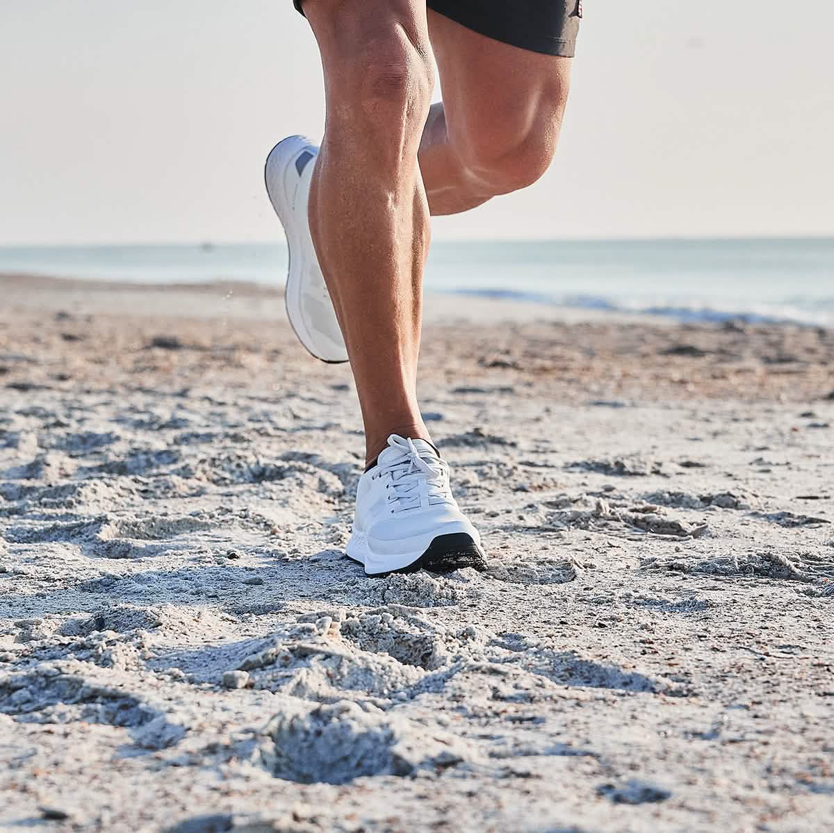 A person jogging on a sandy beach, wearing GORUCK's Rough Runner shoes in light grey and dark grey for extra comfort, paired with black shorts. The ocean is visible in the background under a clear sky.