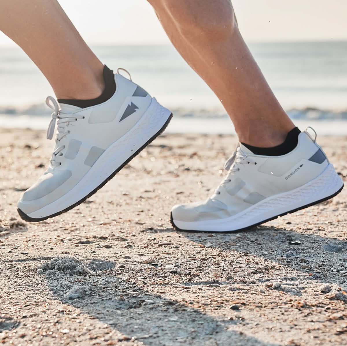Close-up of a person running on a sandy beach, wearing GORUCK's Rough Runner shoes in light grey and dark grey. The background features a blurred view of the ocean under a clear sky.
