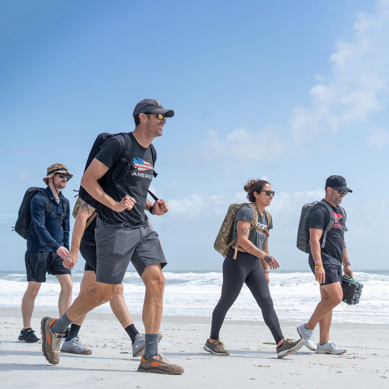 Five people with rucksacks walk together on a sandy beach under a blue sky with scattered clouds, enjoying a day of rucking and the comfort of the Rucker 5.0’s built-in lumbar support.