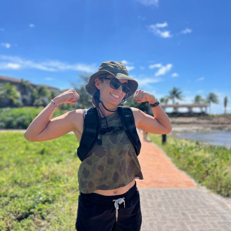 Smiling woman in sunglasses and a hat poses outdoors near the beach on a sunny day, wearing the Spy Ruck
