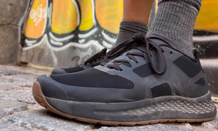Person wearing black sneakers and gray socks, standing on cobblestone with graffiti in the background.